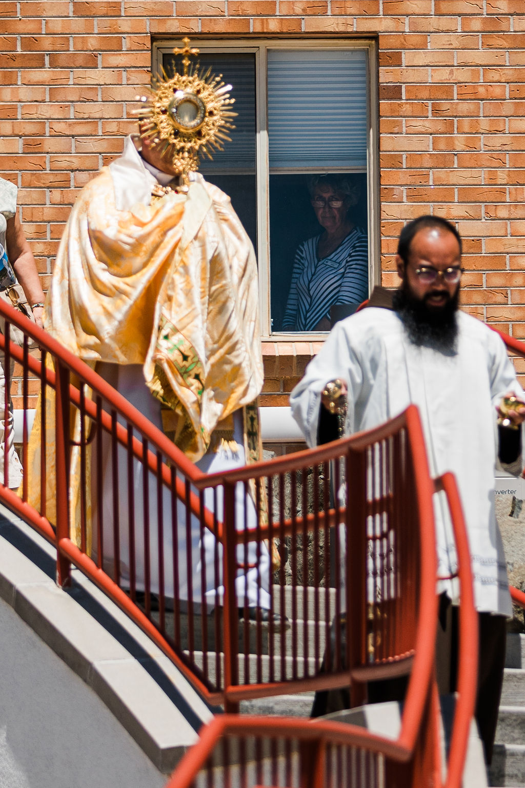 Priest in gold vestments carries a monstrance down red-railed steps, with another cleric in white and a woman watching from a window.