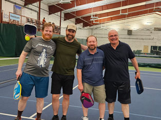 Four men smiling on an indoor tennis court, each holding a pickleball paddle. Background shows nets and banners. Casual mood.