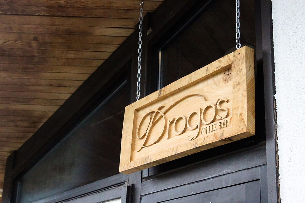 Wooden sign for "Drogo's Coffee Bar" hangs on chains outside a building. The background features a wooden ceiling.