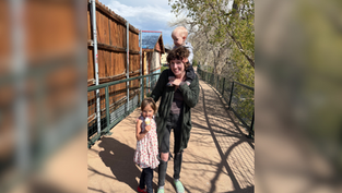 Adult holds toddler on shoulders, standing with a child eating ice cream on a sunny pathway with trees and wooden fence background. Smiling mood.