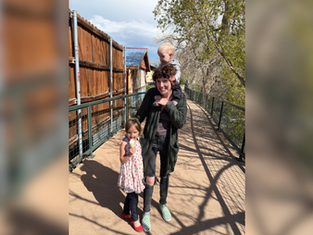 Adult holds toddler on shoulders, standing with a child eating ice cream on a sunny pathway with trees and wooden fence background. Smiling mood.