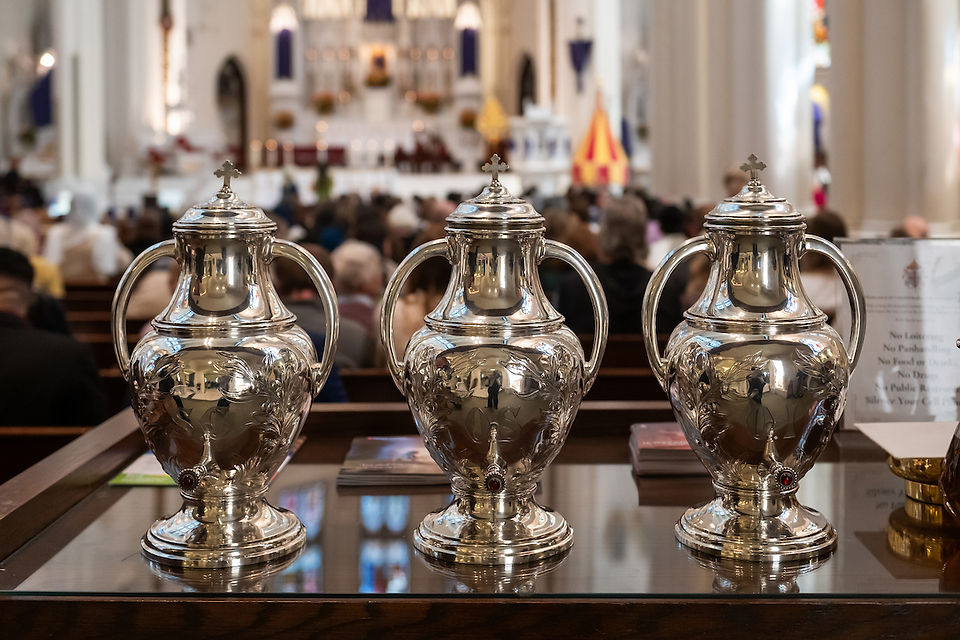 Three ornate silver urns on a table in a crowded church interior. The background shows blurred pews and altar, conveying a solemn mood.