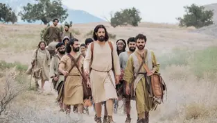 A group of people in historical attire walk through a desert landscape, with mountains in the background. The mood is calm and purposeful.