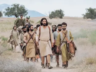 A group of people in historical attire walk through a desert landscape, with mountains in the background. The mood is calm and purposeful.