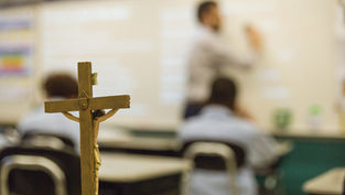 Crucifix in focus in a classroom; blurred teacher writes on whiteboard. Students seated, vibrant wall colors in the background.