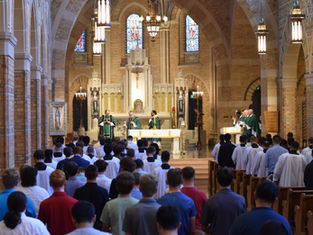 Clergy in white robes process in a grand church with arched ceilings and stained glass. A solemn ceremony unfolds at the ornate altar.