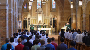 Clergy in white robes process in a grand church with arched ceilings and stained glass. A solemn ceremony unfolds at the ornate altar.