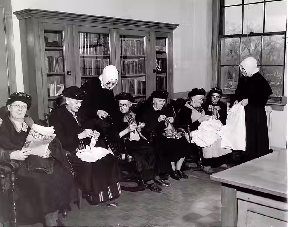 A vintage, black-and-white photo of a group of older women sitting in wooden chairs and knitting or reading. They are all wearing religious habits and head coverings. In the background appears a full bookshelf and a window. The mood is peaceful.