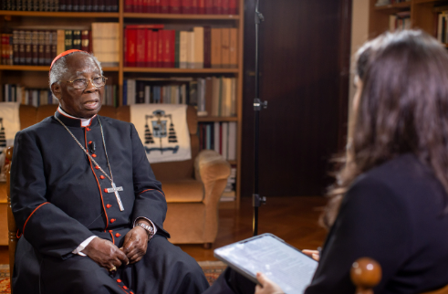 A cardinal in red and black robes sits for an interview with a woman holding a tablet, set in a library with bookshelves and warm lighting.