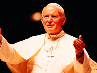 Man in white religious attire gestures expressively, wearing a cross necklace. Dark background, warm lighting, serene expression.