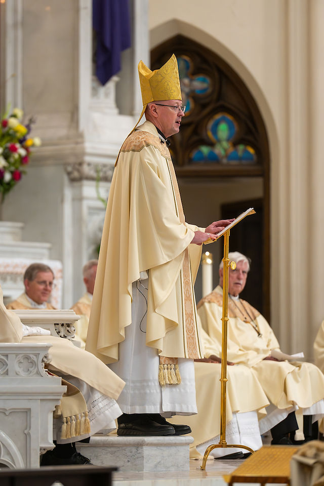 A bishop in a gold robe and mitre speaks at a pulpit in a cathedral with stained glass. Clergy in beige robes sit nearby. Formal setting.