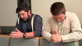 Two men praying with hands clasped in a church, seated on green upholstered chairs. They appear focused and calm. White and blue clothing.