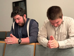 Two men praying with hands clasped in a church, seated on green upholstered chairs. They appear focused and calm. White and blue clothing.