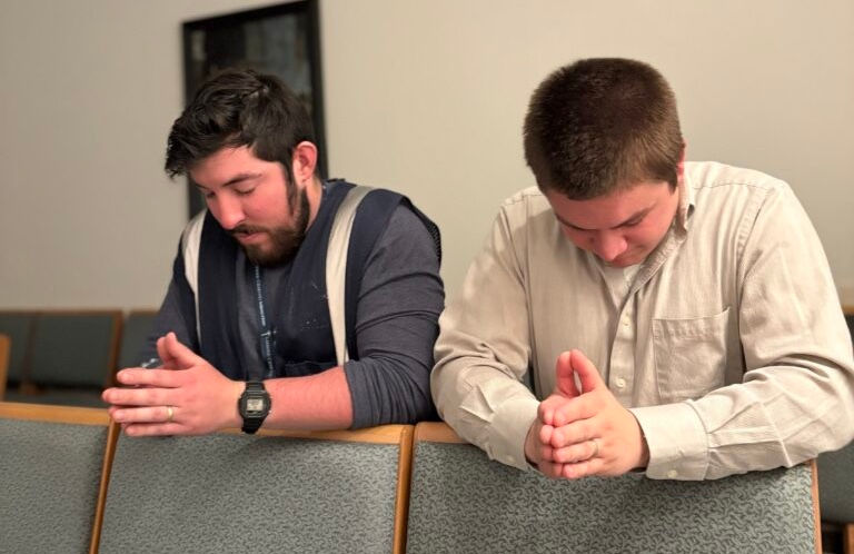 Two men praying with hands clasped in a church, seated on green upholstered chairs. They appear focused and calm. White and blue clothing.