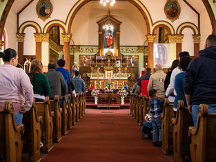 People standing in church pews during a service, facing an ornate altar with statues and floral decorations. The mood is solemn.