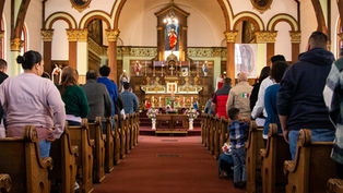 People standing in church pews during a service, facing an ornate altar with statues and floral decorations. The mood is solemn.
