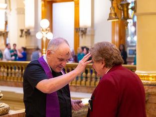 A priest wearing a purple stole imposes ashes on a man in a red blazer's forehead. They stand in an ornate government building, with railings and doorways visible in the background. The mood is prayerful and reflective.