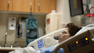 Person in a hospital bed with oxygen tube, eyes closed. Medical equipment and cabinets in the background. Calm, clinical setting.