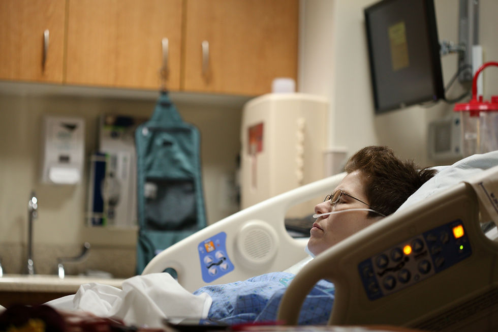 Person in a hospital bed with oxygen tube, eyes closed. Medical equipment and cabinets in the background. Calm, clinical setting.