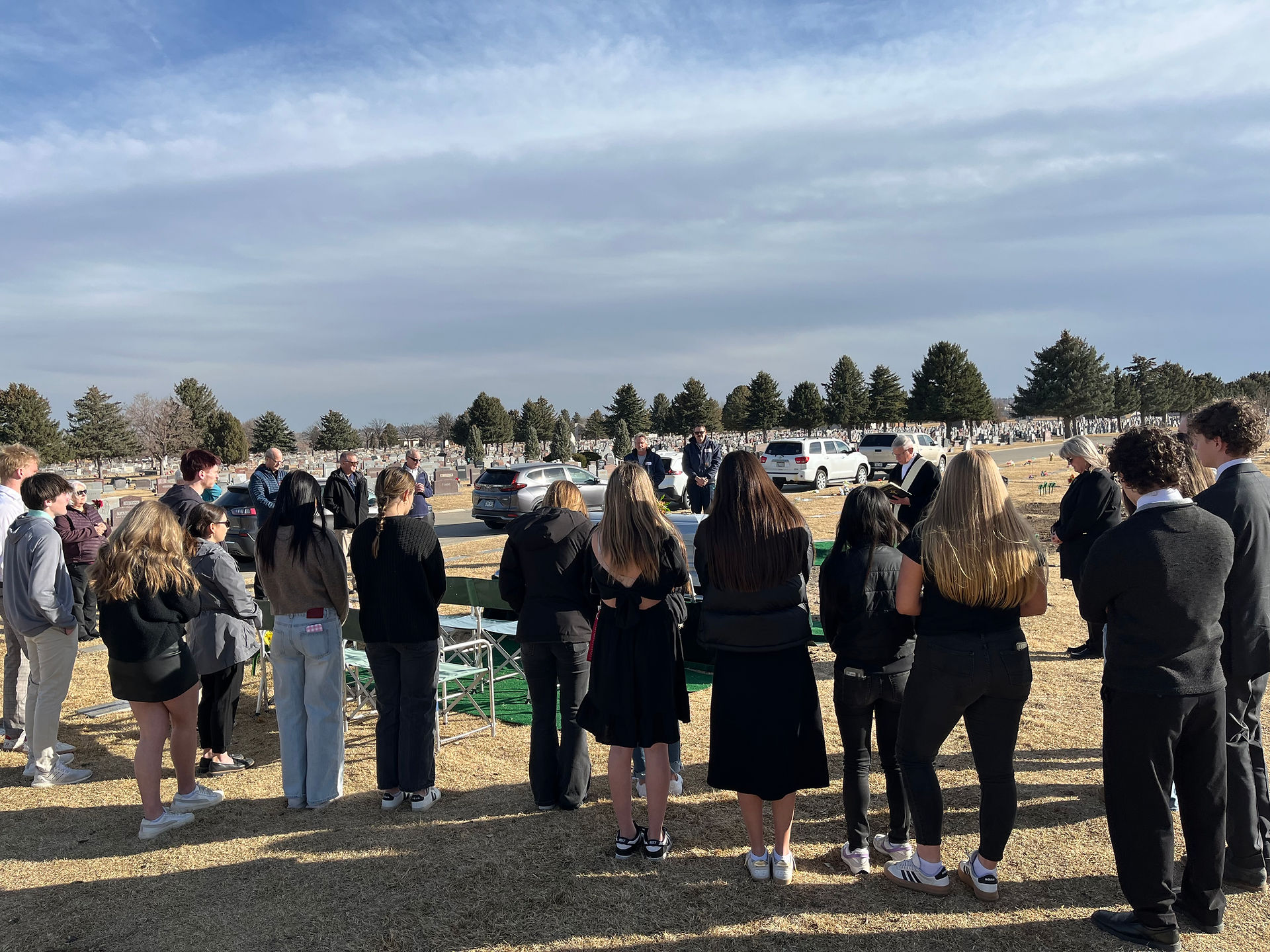A group of people gather for a ceremony in a cemetery. Trees and gravestones are in the background under a partly cloudy sky.