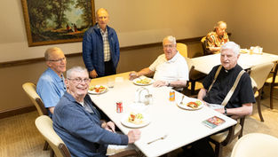 Five elderly men smiling around a dining table with plates of food and drinks. One stands, while others sit. A framed painting decorates the wall.