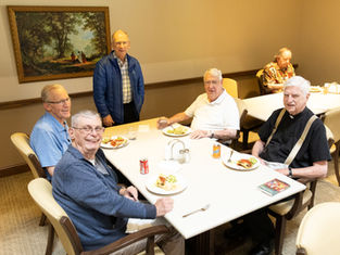Five elderly men smiling around a dining table with plates of food and drinks. One stands, while others sit. A framed painting decorates the wall.