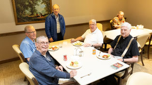 Five elderly men smiling around a dining table with plates of food and drinks. One stands, while others sit. A framed painting decorates the wall.