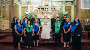A group of people in a church setting wearing black attire with blue and green sashes, standing in front of an ornate altar and wood paneling.