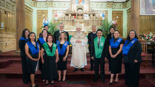 A group of people in a church setting wearing black attire with blue and green sashes, standing in front of an ornate altar and wood paneling.