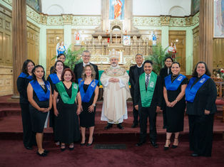 A group of people in a church setting wearing black attire with blue and green sashes, standing in front of an ornate altar and wood paneling.