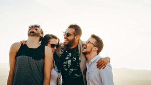 Four friends laugh together outdoors, wearing casual clothes and sunglasses, against a bright sky. Warm, joyful mood.