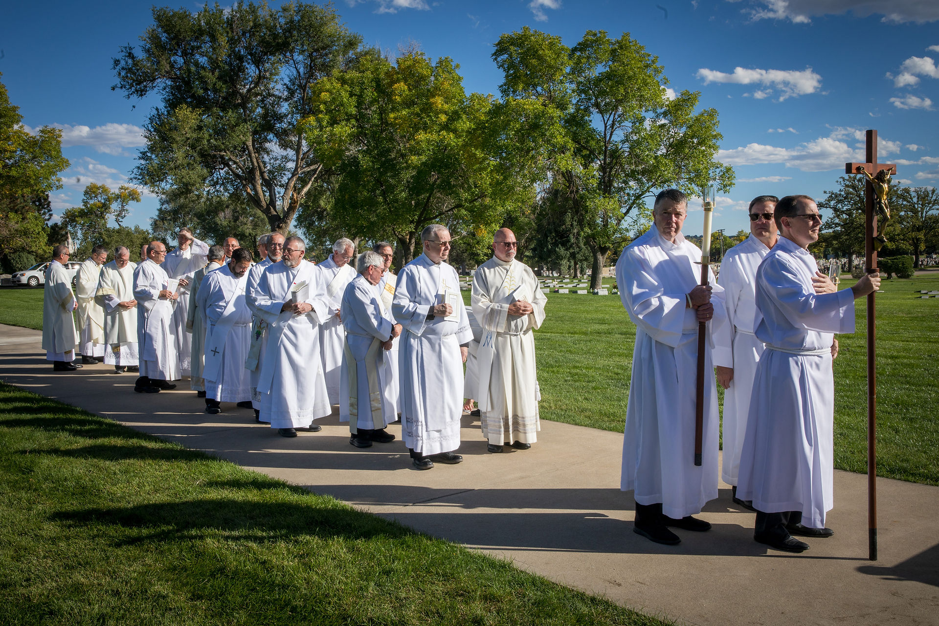 Procession of priests in white robes holding crosses and candles, walking on a sunny path in a grassy park with trees and blue sky.