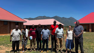 A group of ten people stands outside with red-roofed buildings and mountains in the background, under a clear blue sky, smiling at the camera.