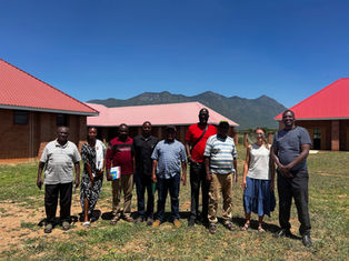 A group of ten people stands outside with red-roofed buildings and mountains in the background, under a clear blue sky, smiling at the camera.