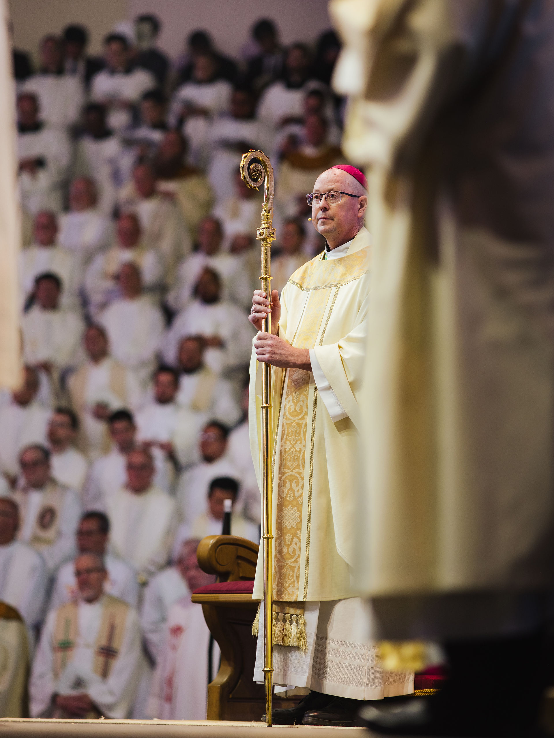 Bishop in ornate robes holds a golden crozier, standing solemnly among a crowd of robed clergy in a church setting.