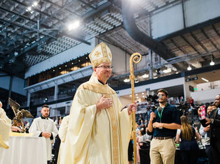 A bishop in ornate robes and hat holds a staff in a large indoor venue, surrounded by clergymen, photographers, and attendees.