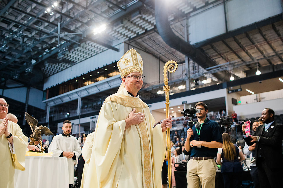 A bishop in ornate robes and hat holds a staff in a large indoor venue, surrounded by clergymen, photographers, and attendees.