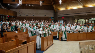 Clergy in white robes with green stoles stand and pray in a wooden church interior. Latin text on walls, solemn atmosphere.