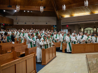 Clergy in white robes with green stoles stand and pray in a wooden church interior. Latin text on walls, solemn atmosphere.