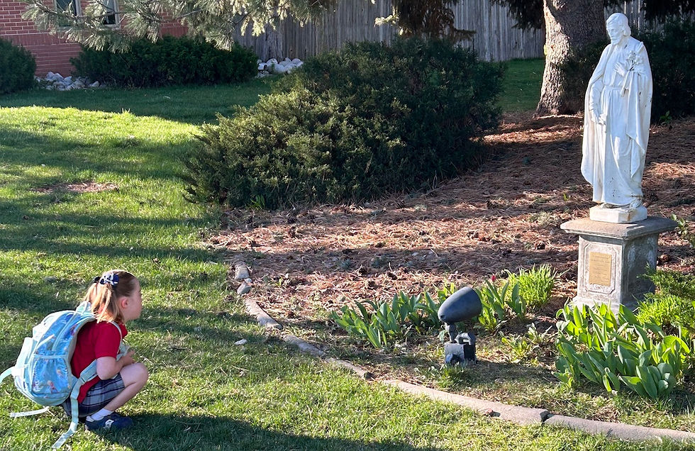 Child in red shirt with blue backpack squats on grass, gazing at a white statue of St. Joseph in a garden. Sunny day, trees and bushes in background.