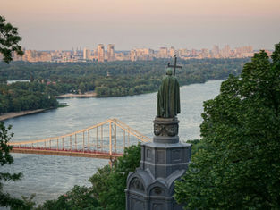 Statue on a hill overlooks a river, bridge, and distant city skyline framed by trees at sunset.
