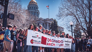 A group holds a Colorado March for Life banner outside a domed building, with trees and a U.S. flag in the background. Sunny day.