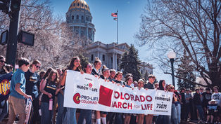A group holds a Colorado March for Life banner outside a domed building, with trees and a U.S. flag in the background. Sunny day.