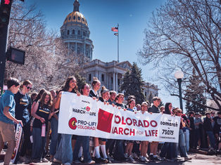 A group holds a Colorado March for Life banner outside a domed building, with trees and a U.S. flag in the background. Sunny day.