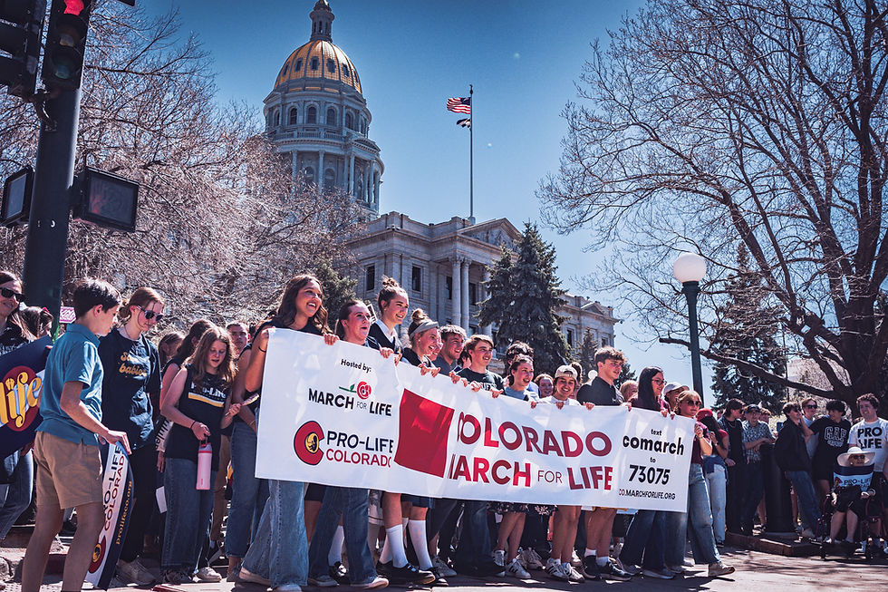 A group holds a Colorado March for Life banner outside a domed building, with trees and a U.S. flag in the background. Sunny day.