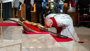 Bishop in white and red robes kisses a crucifix on red cloth in a church, with attendees watching. Marble floor reflects the scene.