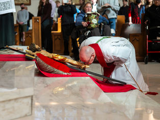 Bishop in white and red robes kisses a crucifix on red cloth in a church, with attendees watching. Marble floor reflects the scene.