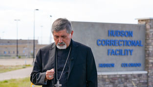 Bishop in black holds a rosary outside Hudson Correctional Facility sign in Hudson, Colorado, looking somber.
