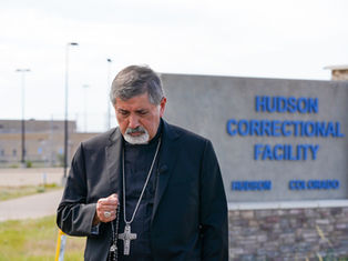 Bishop in black holds a rosary outside Hudson Correctional Facility sign in Hudson, Colorado, looking somber.