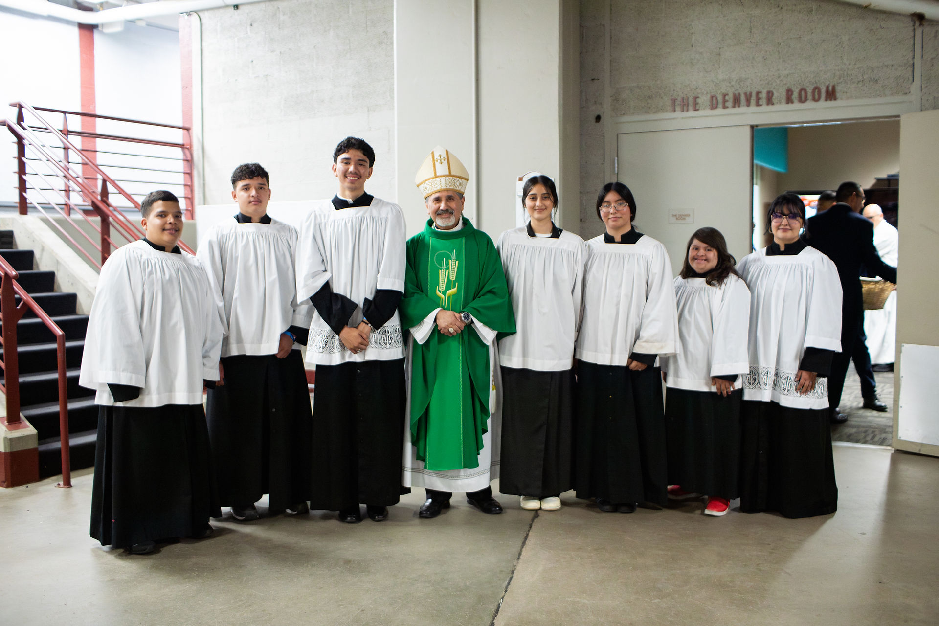 A group of altar servers in white albs and black cassocks stand smiling beside a bishop dressed in green liturgical vestments. They are posed in a hallway near a door labeled “The Denver Room,” likely before or after a Mass. The bishop stands at the center, and the setting is informal but reverent.